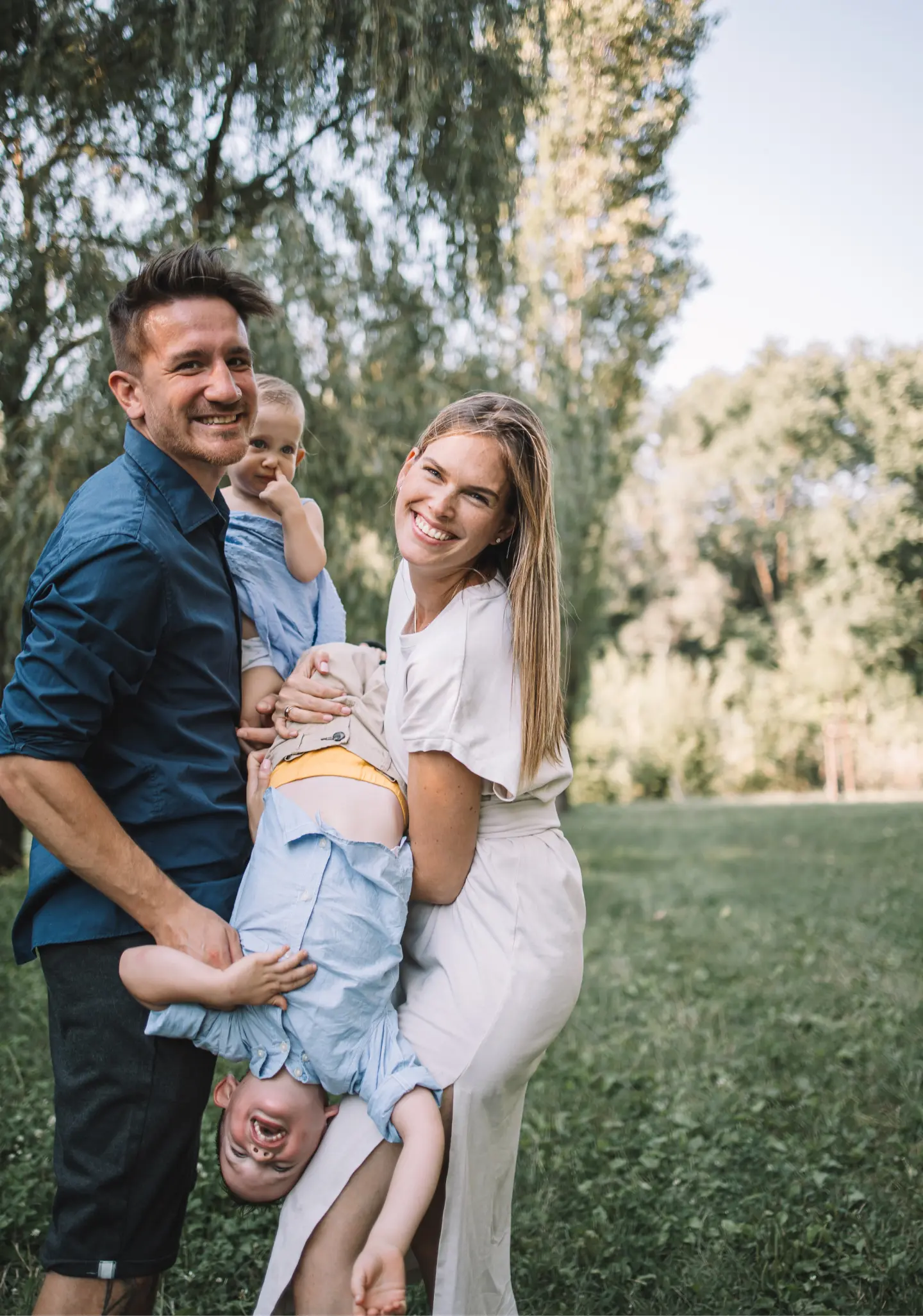 anyuka és apuka a kisfiukkal és a kislányukkal mosolyog a kamerába, az kisfiút fejjel lefelé lógatják, családi fotózás