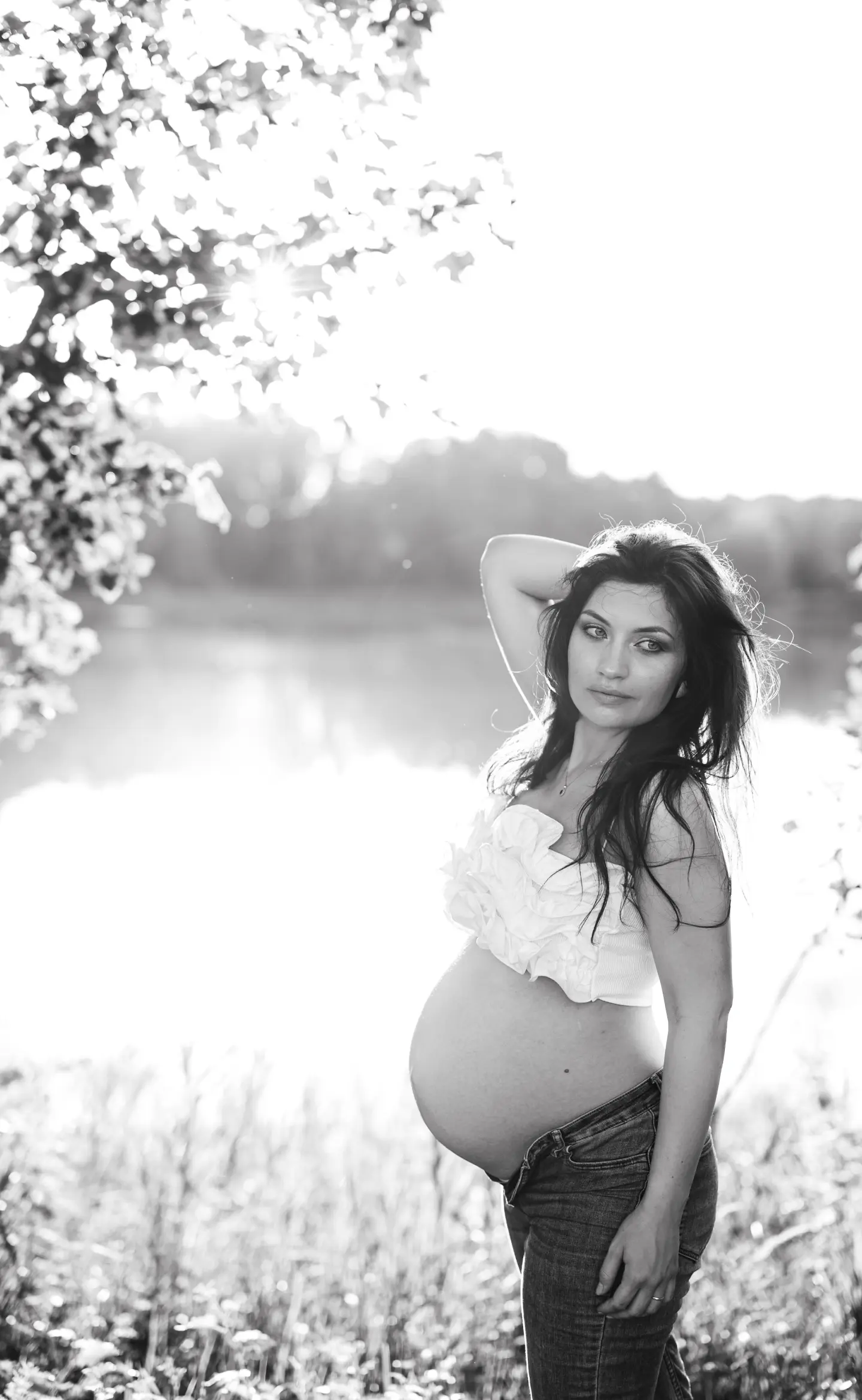 Pocakos nő nyári fényben, tóparton állva, keze a hasán