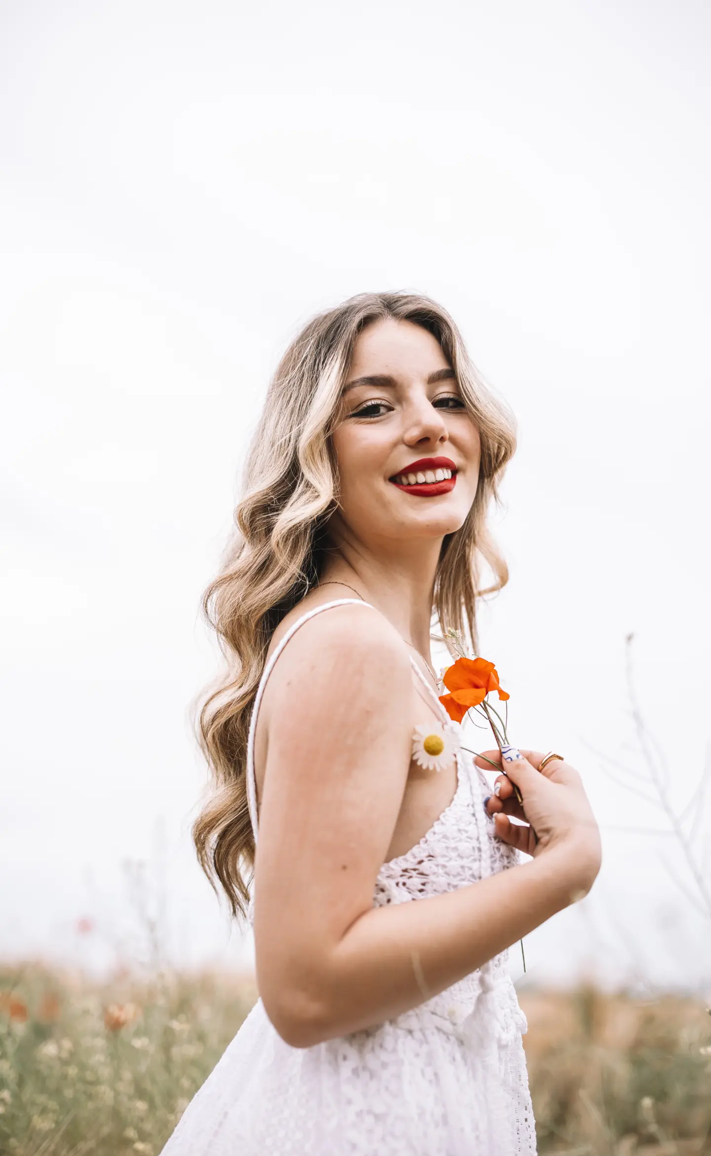 egy hosszú szőke hajú, fehér ruhás lány egy 2 virágszálat a kezében tartva mosolyog a kamerába, portré fotózás