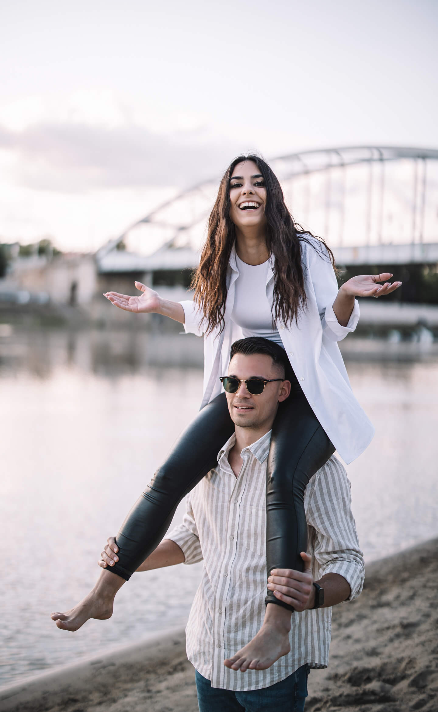 Lány nevetve ül a barátja vállán vízparton, páros lifestyle fotózás