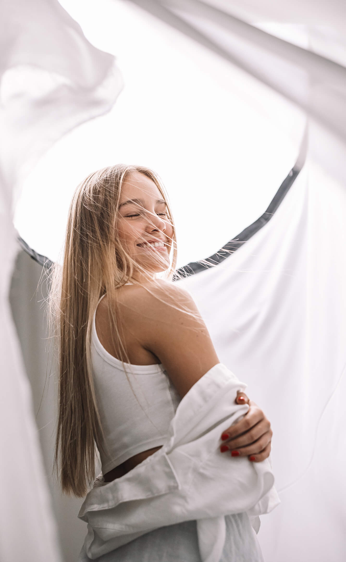 Fiatal nő nevetve fordul hátra, szél fújja a haját, világos háttér előtt